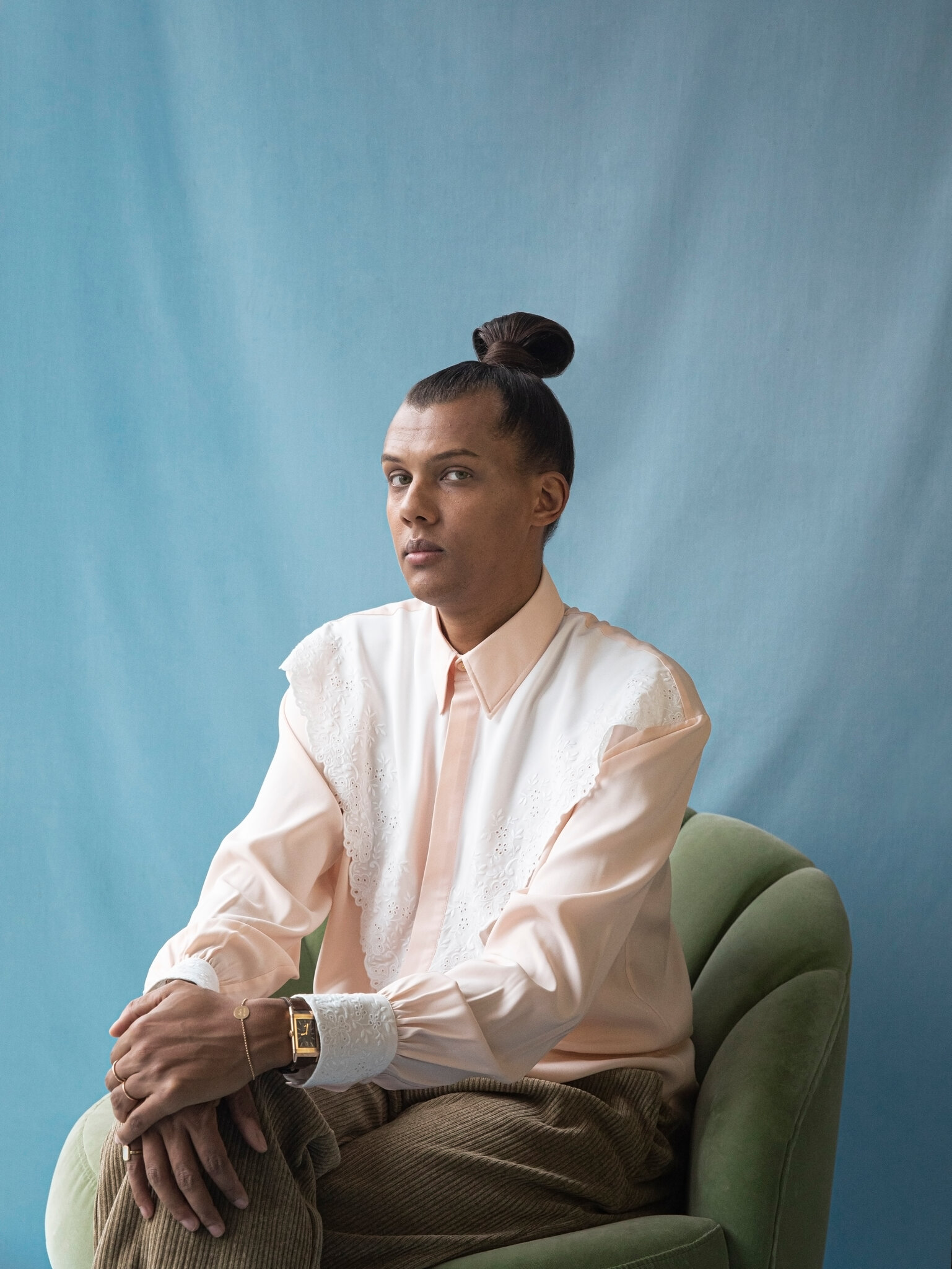 Stromae seated in a green velvet chair, peach blouse, blue backdrop